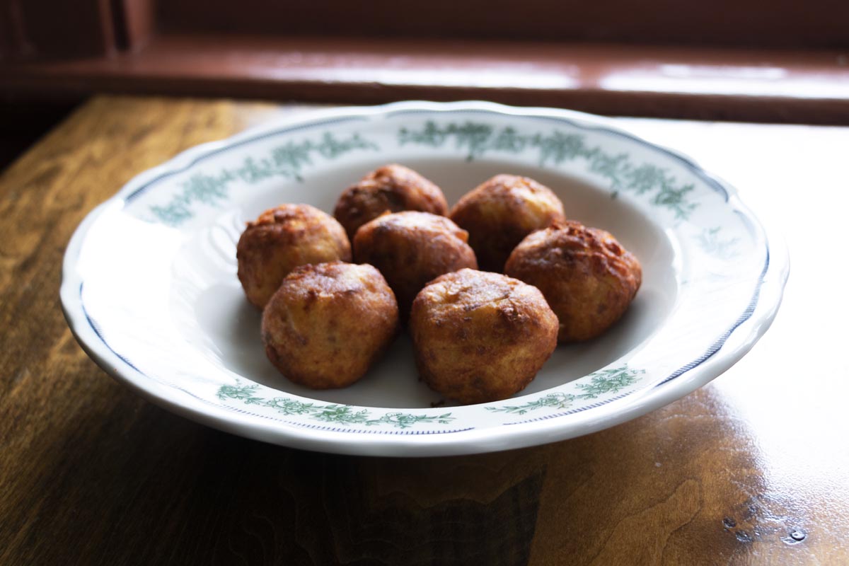 Eagle Tavern’s Pork and Potato Balls Shallow white bowl with green floral pattern around rim, filled with fried meatballs, sitting on wooden table