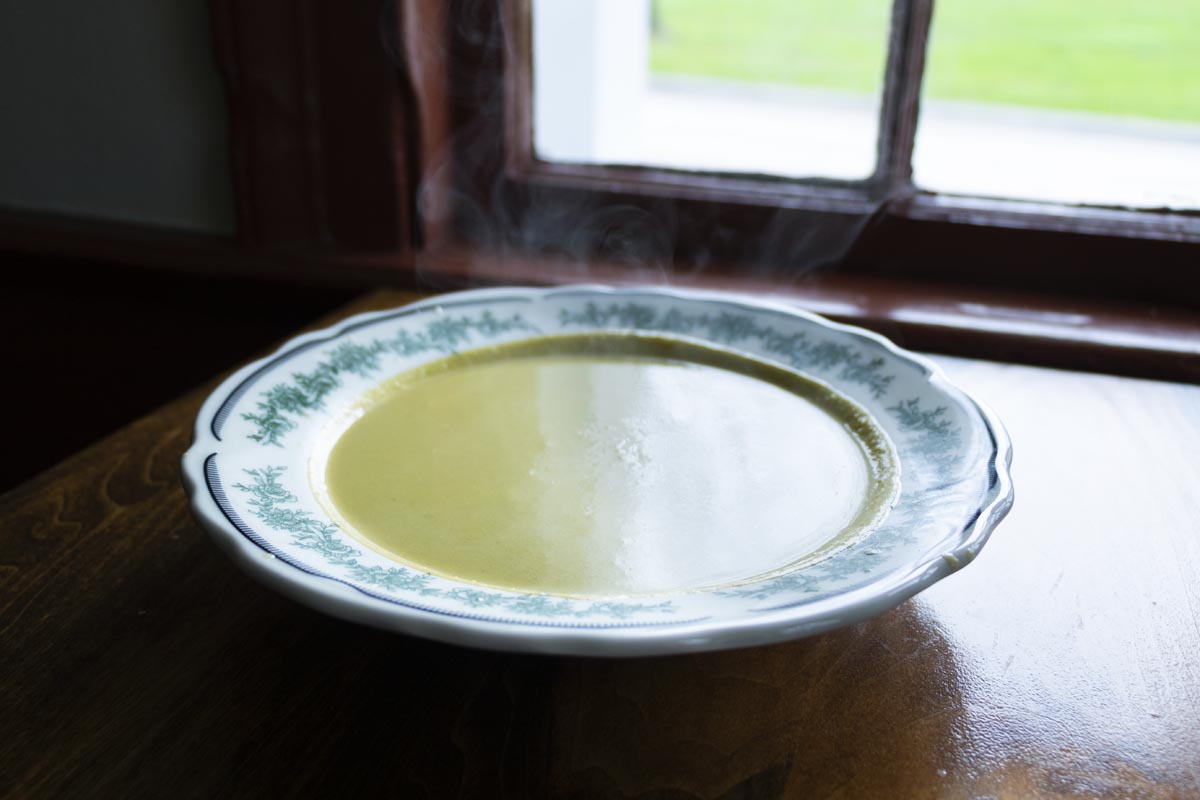 Eagle Tavern's Asparagus Soup Shallow white bowl with green floral pattern filled with steaming pea-colored soup, sitting on wooden table in front of window