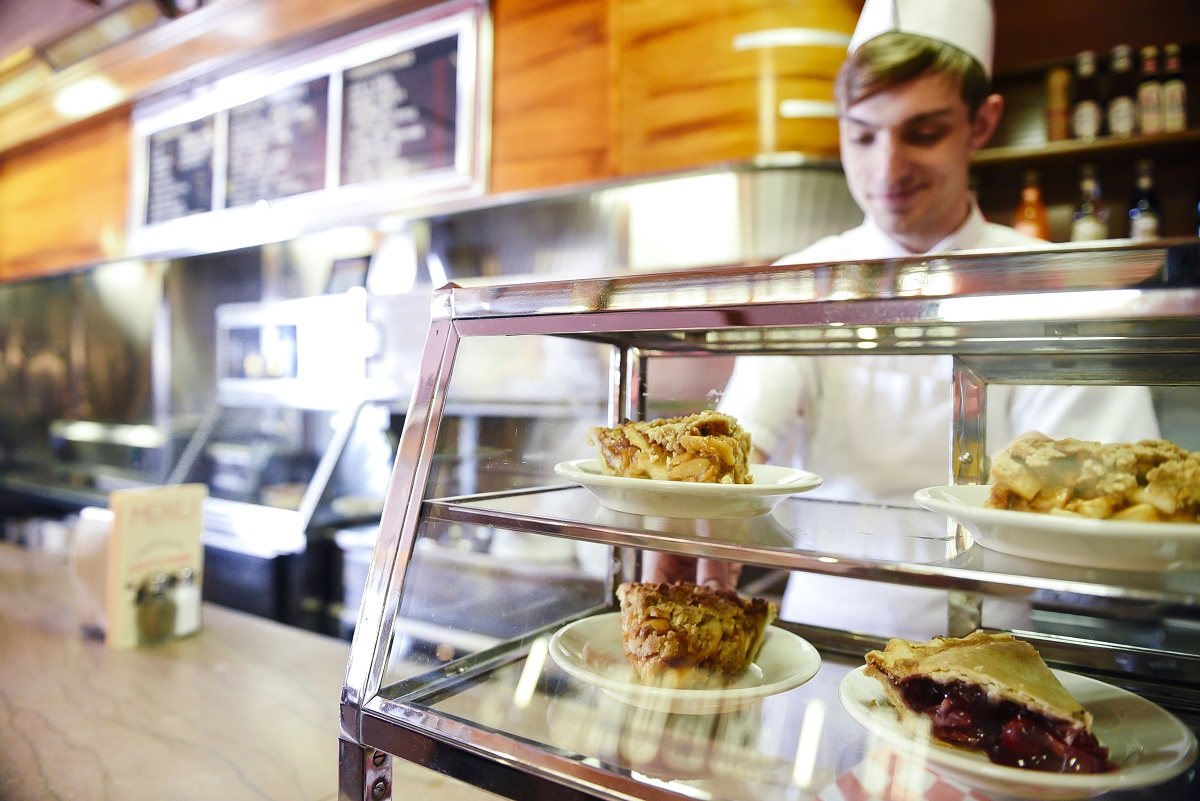 A glass case filled with slices of pie sits on a lunch counter with a person in a white cap and and apron behind the counter 