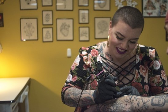 Smiling person in black gloves tattoos another person's arm; framed art hangs on a wall behind them